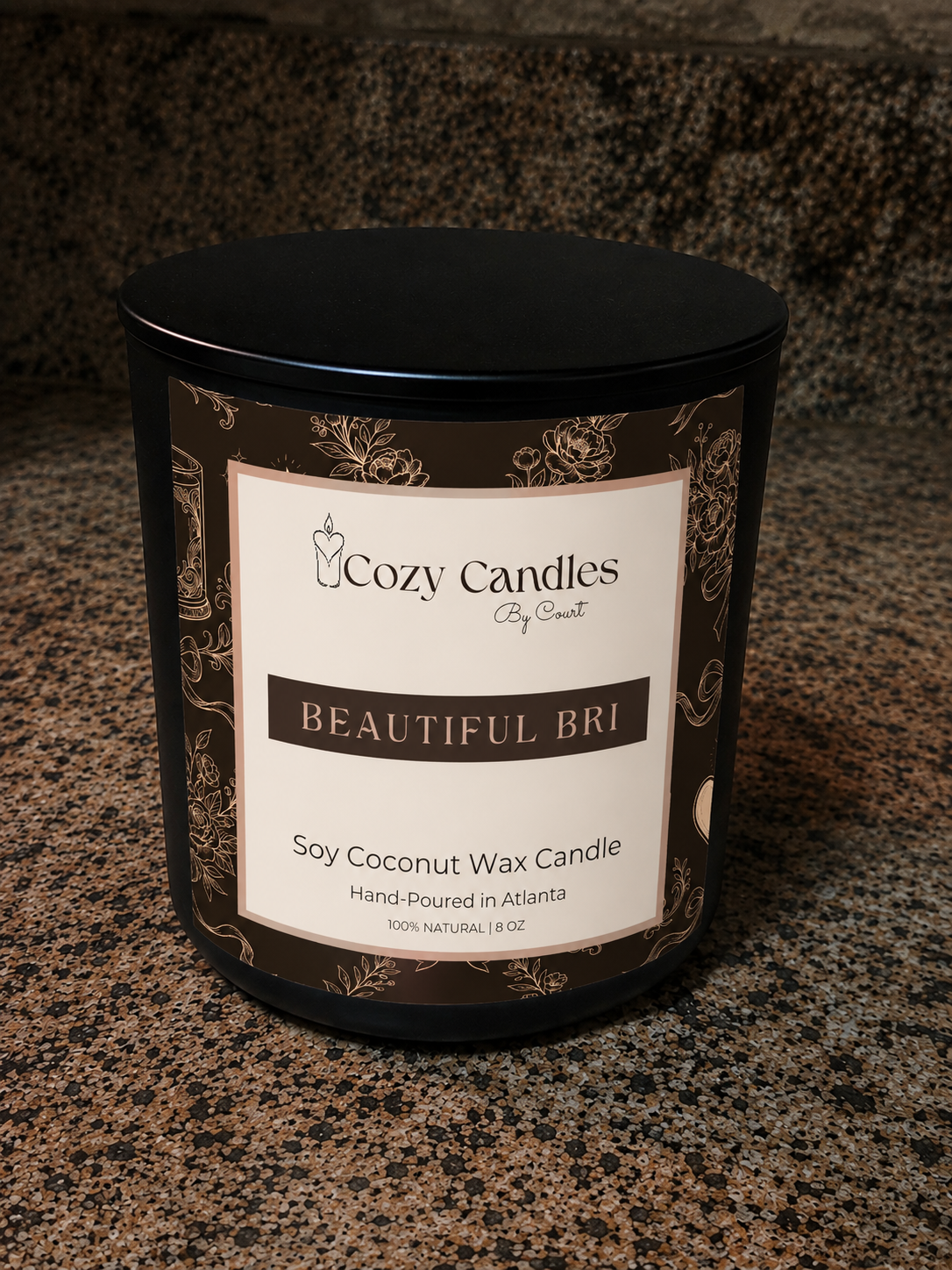 A black soy coconut wax candle by Cozy Candles, titled 'Beautiful Bri', shown on a speckled granite countertop. The candle is in a black metal tin with a cream and dark brown label featuring ornate gold-toned scrollwork.