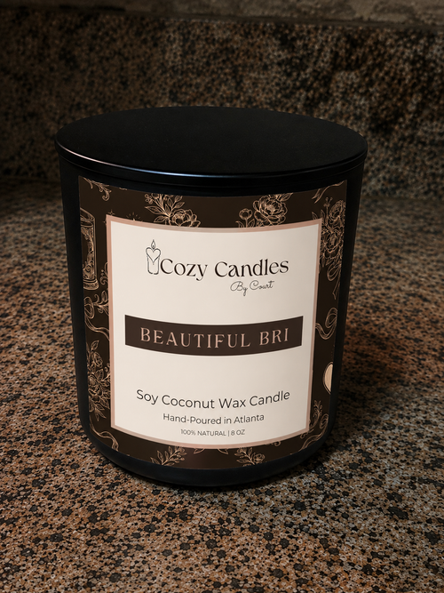 A black soy coconut wax candle by Cozy Candles, titled 'Beautiful Bri', shown on a speckled granite countertop. The candle is in a black metal tin with a cream and dark brown label featuring ornate gold-toned scrollwork.