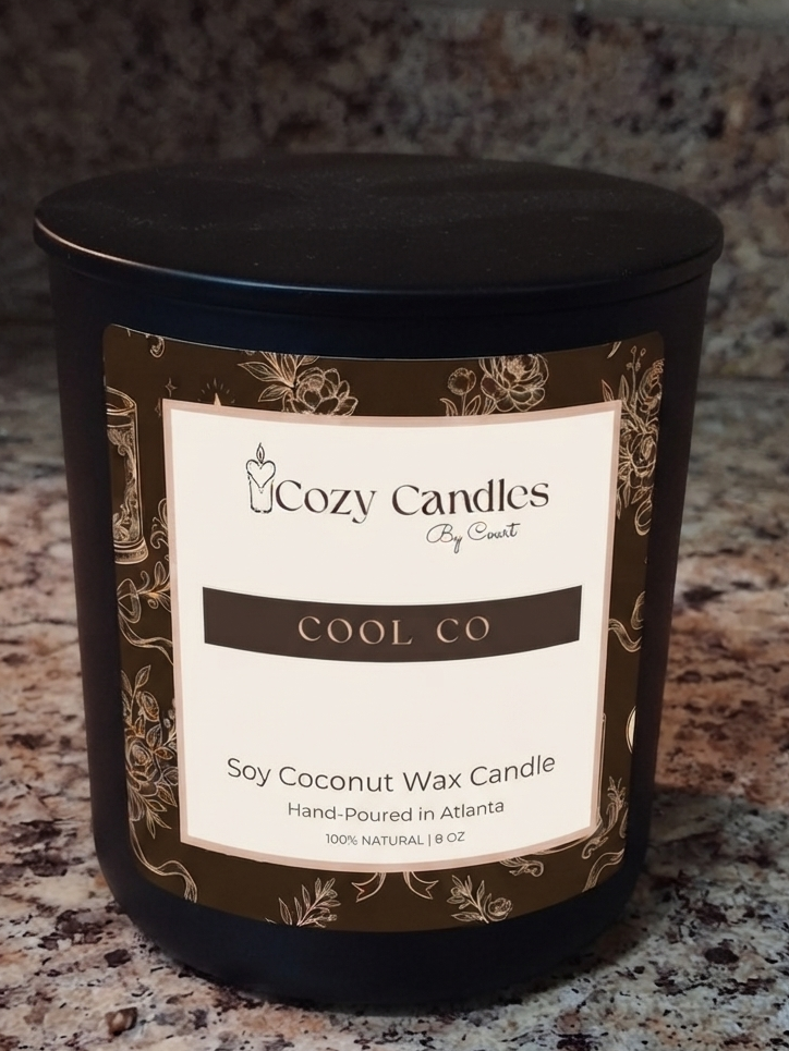 A black glass jar candle with a matching lid and a cream label reading 'Cozy Candles by Scents COOL CO Soy Coconut Wax Candle' on a speckled granite countertop.