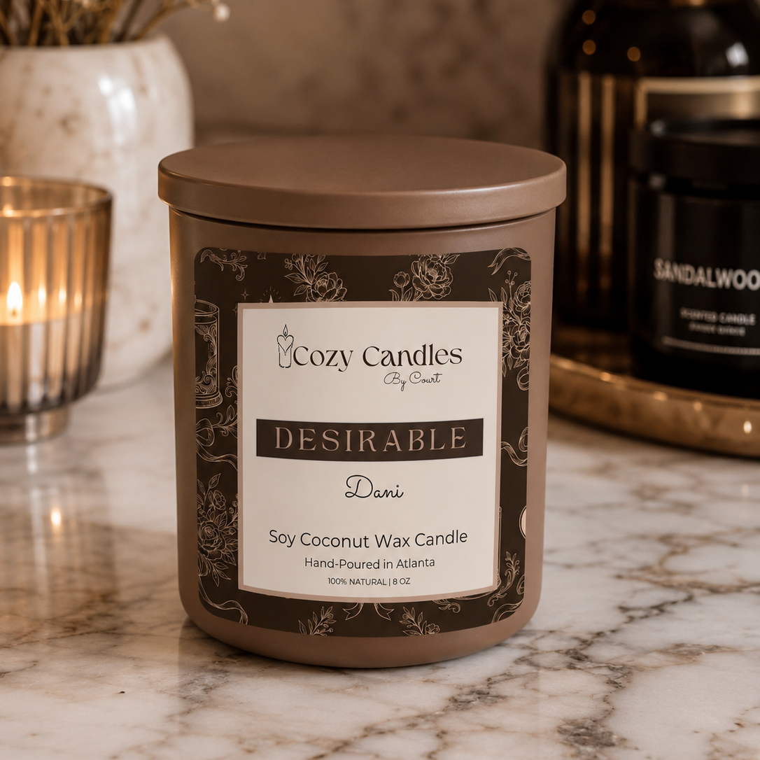 A Cozy Candles 'Desirable' Dani scented soy coconut wax candle in a brown metal tin with a cream label, styled on a marble countertop with a lit candle and other home decor in the background.