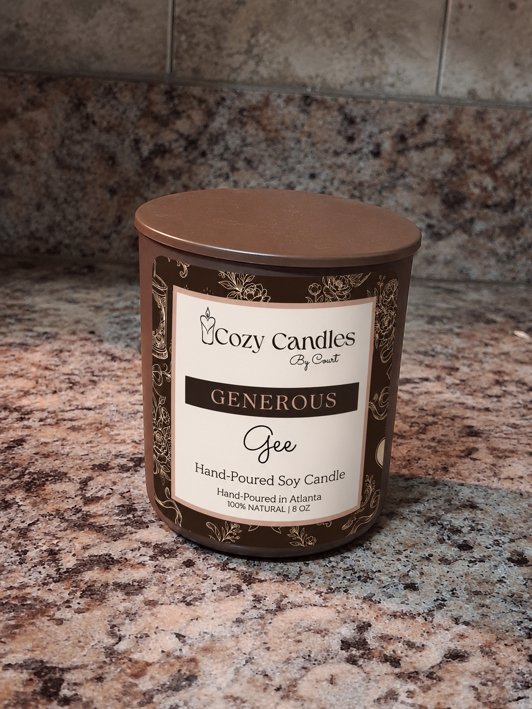 A Cozy soy candle in a brown glass jar with a matching brown lid and a cream label reading 'Generous Gee'. The candle is placed on a speckled granite countertop with a tiled backsplash behind it.
