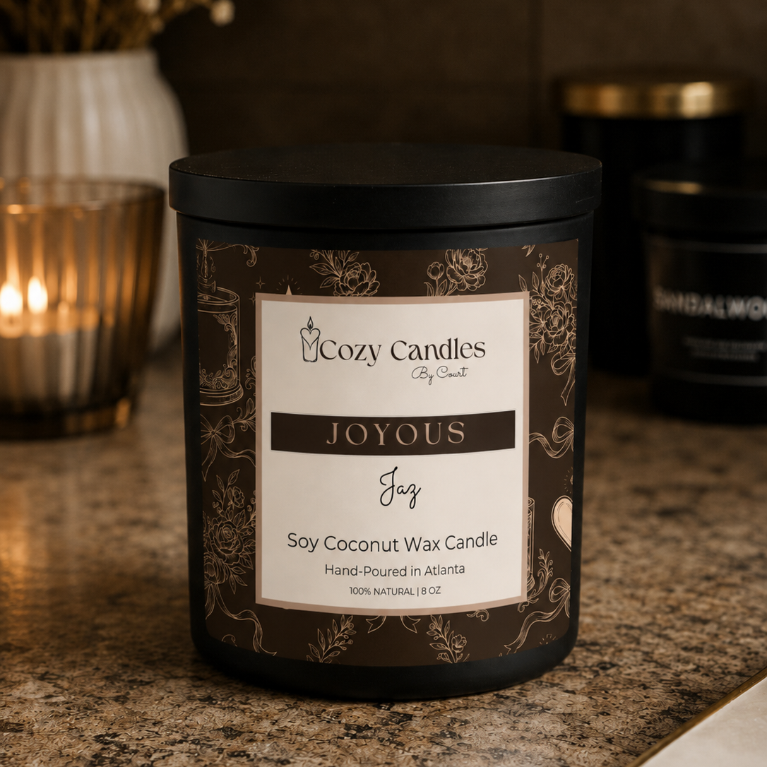 A Cozy Candles 'Joyous Joy' soy coconut wax candle in a dark brown decorative jar with a black lid, set on a granite countertop with a warm lit candle and dried flowers in the background.